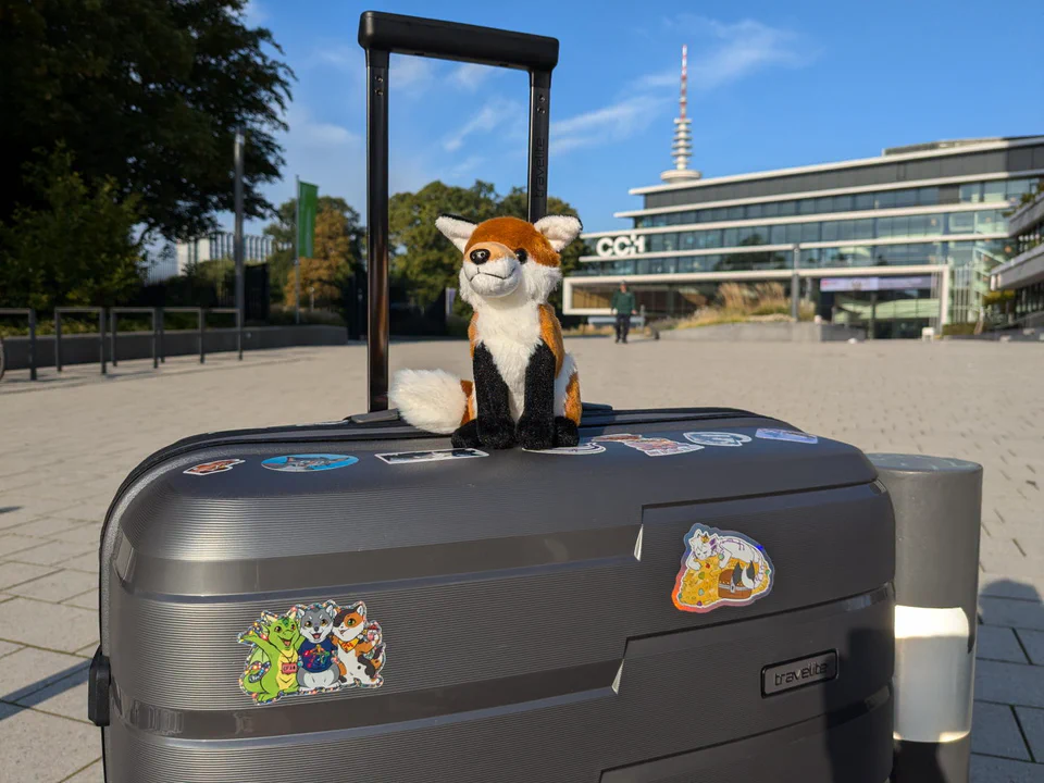 Photo of a little red fox plush sitting on a large gray travel suitcase covered in furry stickers, with the Congress Center Hamburg in the background