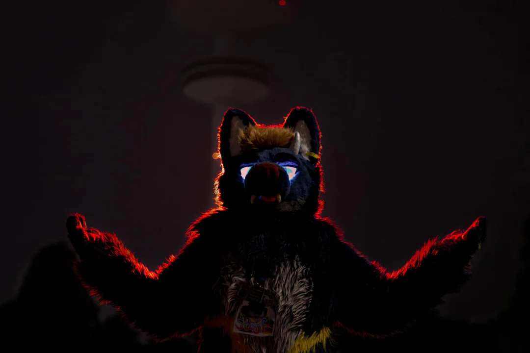 Close-up photo of a dragon-wolf fursuiter with blue fur and glowing blue eyes, arms raised in a powerful pose, with the large Hamburg TV tower in the background