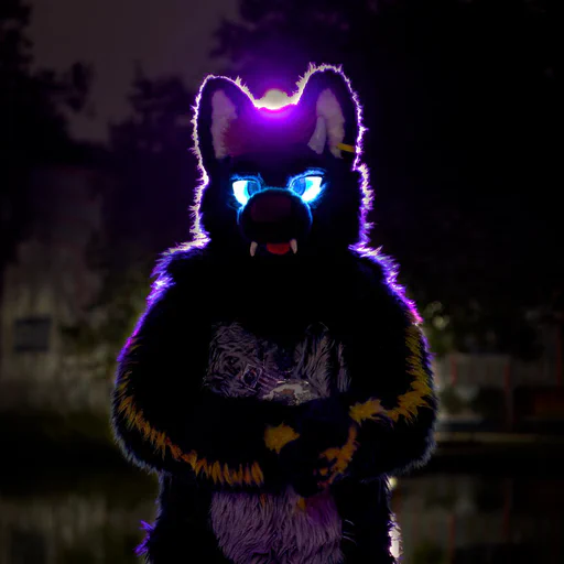 Photo of a dragon-wolf fursuiter with glowing blue eyes, rubbing his hands together while looking at the camera. He is lit with purple backlight.