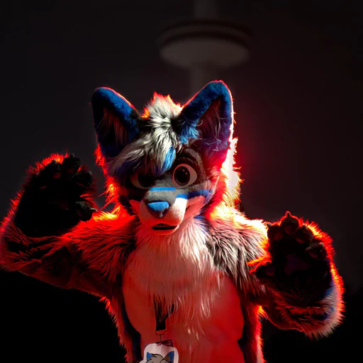 Close-up photo of a wusky fursuiter with light gray fur and blue stripes, both hands raised as if to pounce on the camera, lit with a red back light with a TV tower out of focus in the background.