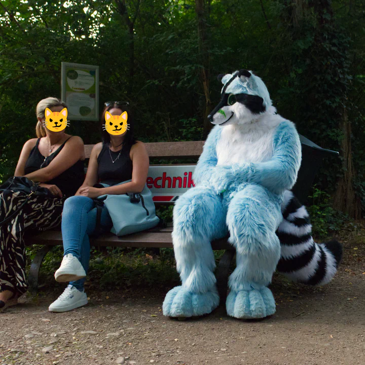 A blue raccoon fursuiter sitting on a wooden bench next to two guests