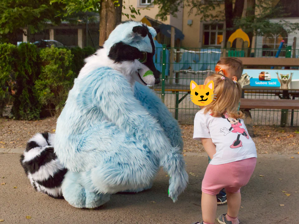 A blue raccoon fursuiter squatting on the ground, with two children looking at him excitedly