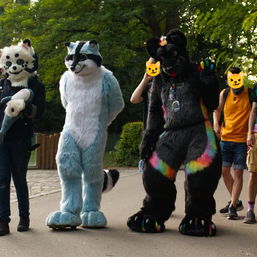 Group photo of three fursuiters (a black and white demon dog holding a plush shark, a blue raccoon, and a black jaguar with rainbow stripes), surrounded by five friends without fursuits. They're standing on a stone walkway inside a zoo, with an outdoor enclosure in the background.
