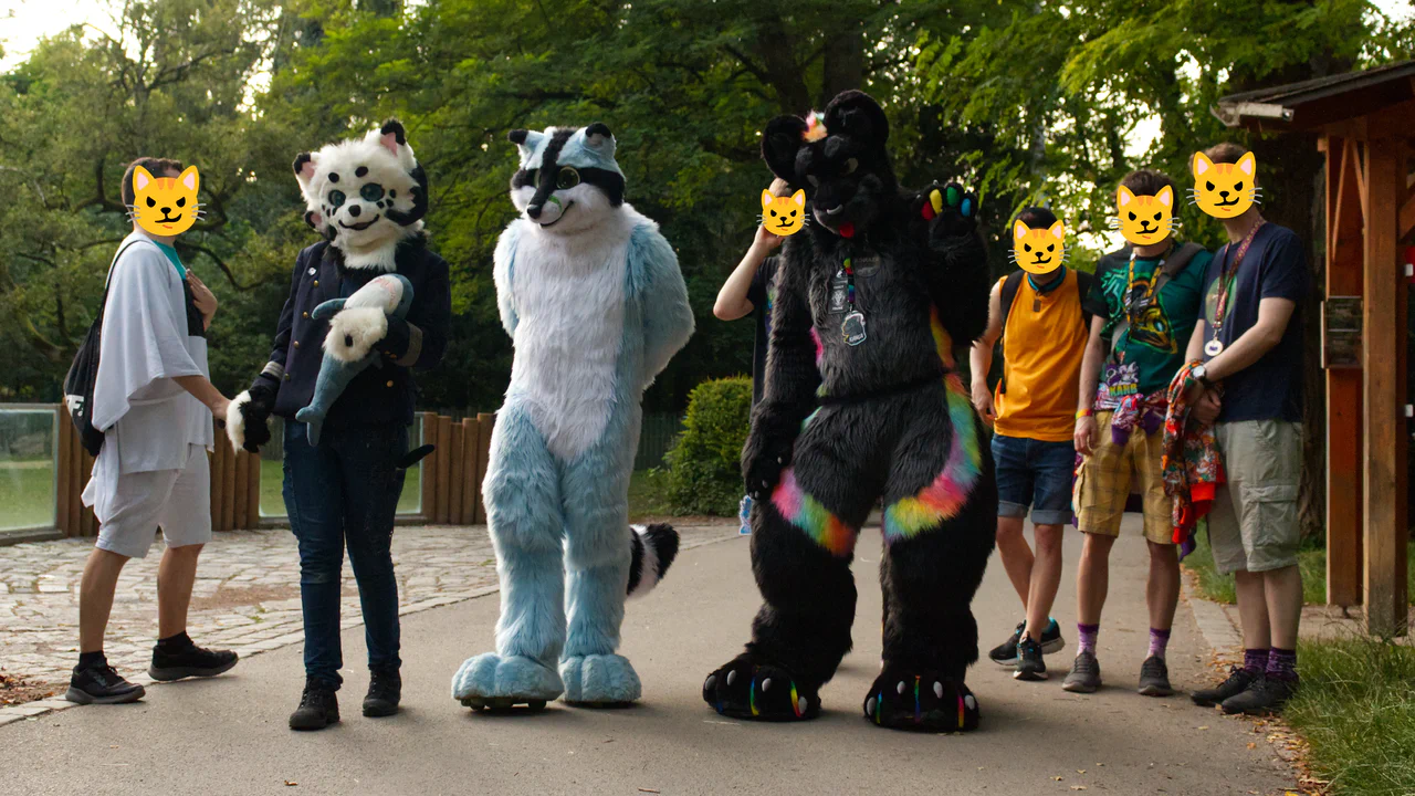 Group photo of three fursuiters (a black and white demon dog holding a plush shark, a blue raccoon, and a black jaguar with rainbow stripes), surrounded by five friends without fursuits. They're standing on a stone walkway inside a zoo, with an outdoor enclosure in the background.
