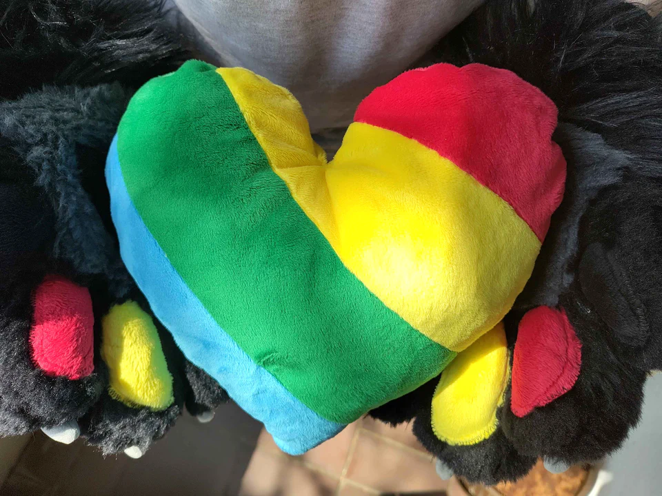 Photo of a plush heart made of short minky fabric in four diagonal stripes: red, yellow, green, and blue. I am holding it in my fursuit paws.
