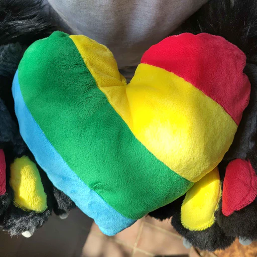 Photo of a plush heart made of short minky fabric in four diagonal stripes: red, yellow, green, and blue. I am holding it in my fursuit paws.
