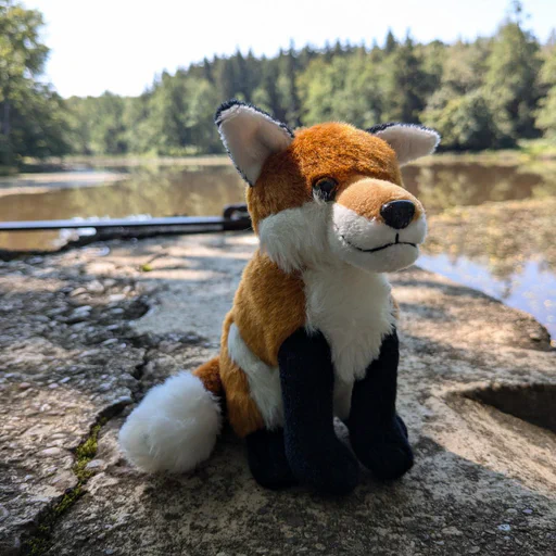 Photo of a red fox plush sitting on a concrete wall overlooking a larger pond in a forest.