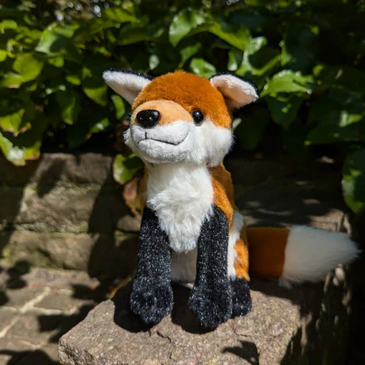 Close-up photo of a small red fox plush, sitting on a stone slab in front of green plants.
