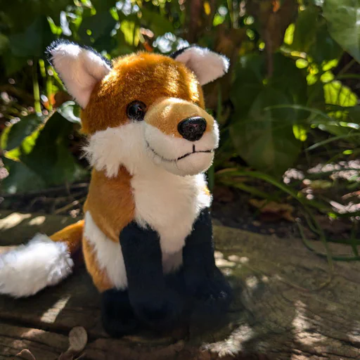 Photo of a small red fox plush, sitting on an old piece of weathered wood with more plants in the background.
