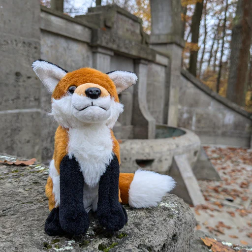 Close-up photo of a small red fox plush, sitting on outer edge of a large stone fountain, currently dry. Everything is covered in autumn leaves, with parts of a forest visible in the background.
