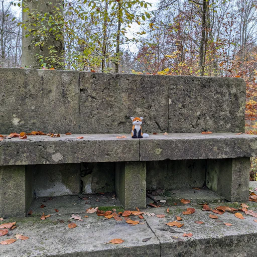 Photo of a small red fox plush sitting on a rather massive stone bench in the middle of a forest, with autumn leaves all around him.
