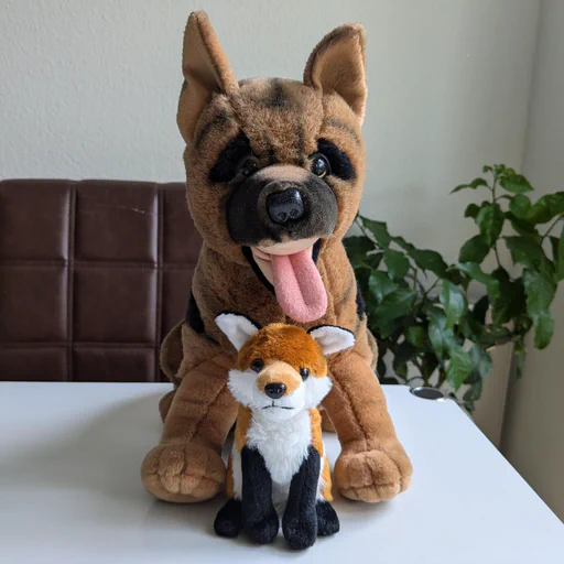 Photo of a small red fox plush sitting on a white living room table, which a much larger doggo plushie behind him. Both are looking straight at the camera.
