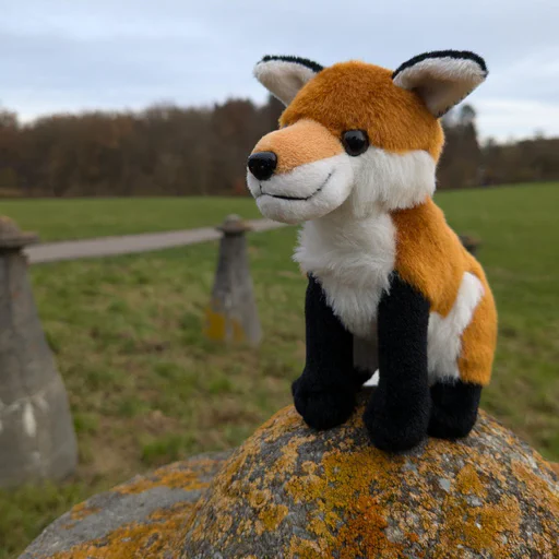 Photo of a small red fox plush sitting on a stone pedestal covered in orange lichen, with more pedestals on a large green field in the background.
