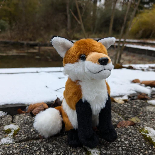 Photo of a small red fox plush sitting on a stone walkway partially covered in snow, with a pond and long grass in the background.
