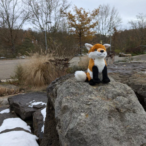 Photo of a small red fox plush sitting on a large stone that's part of a spring, with an empty street and trees in the background.
