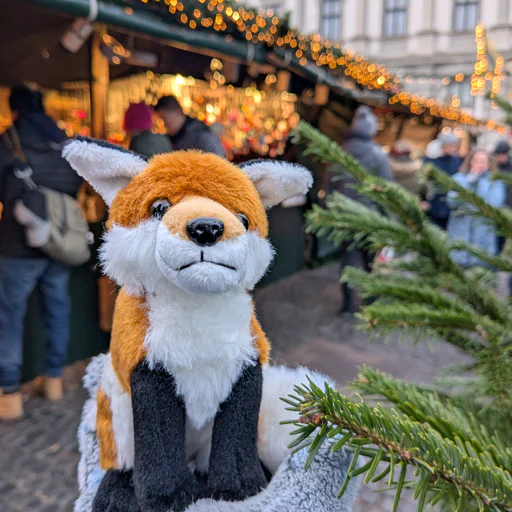 Close-up photo of a small red fox plush, held in a gloved hand next to a Christmas tree on the Augsburg town square. Out of focus in the background, there are people passing by and browsing various booths at the Christmas market
