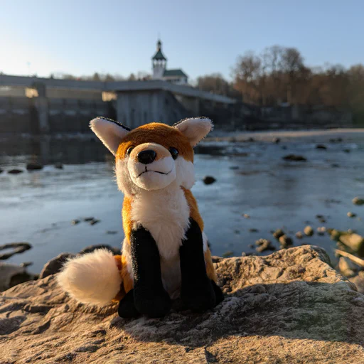 Photo of a small red fox plush sitting on a large stone, with a shallow river and a large weir and bridge structure in the background.
