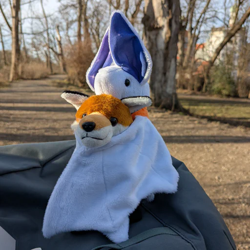 Photo of a white bat plush with purple ears hugging a smaller red fox plush. They're sitting on a gray backpack, with a sunny park out of focus in the background.
